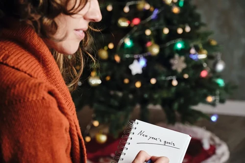 A woman makes a list of New Year’s goals on a notepad while sitting in front of a lit Christmas tree. 
